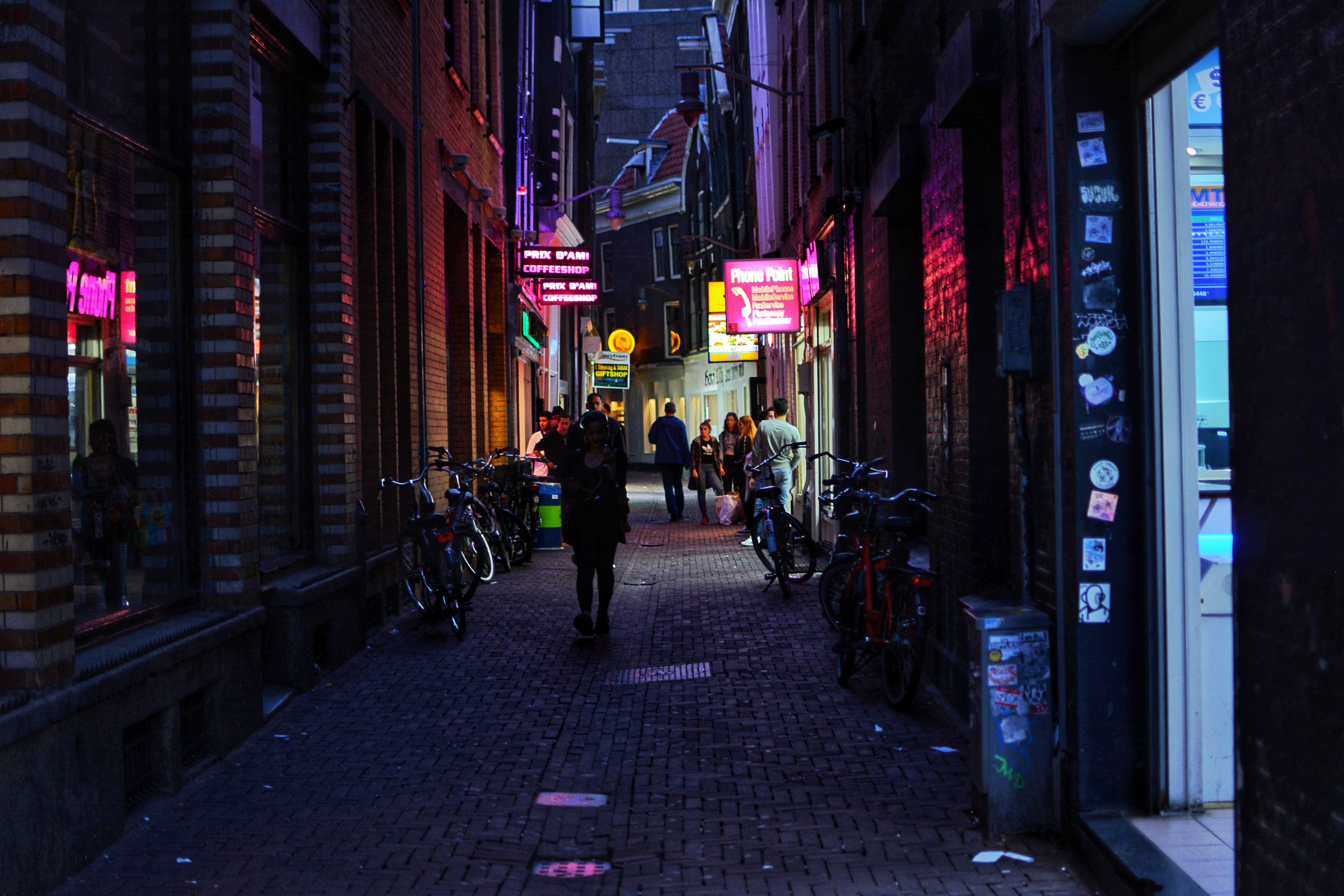 A photograph of a narrow street in Amsterdam, Netherlands at night. Neon signs line the cobblestone street indicating coffee shops, smoke shops, and a public mobile phone among others. Bikes line the edges of the street, and pedestrians walk and stand to talk to each other.A photograph of a narrow street in Amsterdam, Netherlands at night. Neon signs line the cobblestone street indicating coffee shops, smoke shops, and a public mobile phone among others. Bikes line the edges of the street, and pedestrians walk and stand to talk to each other.