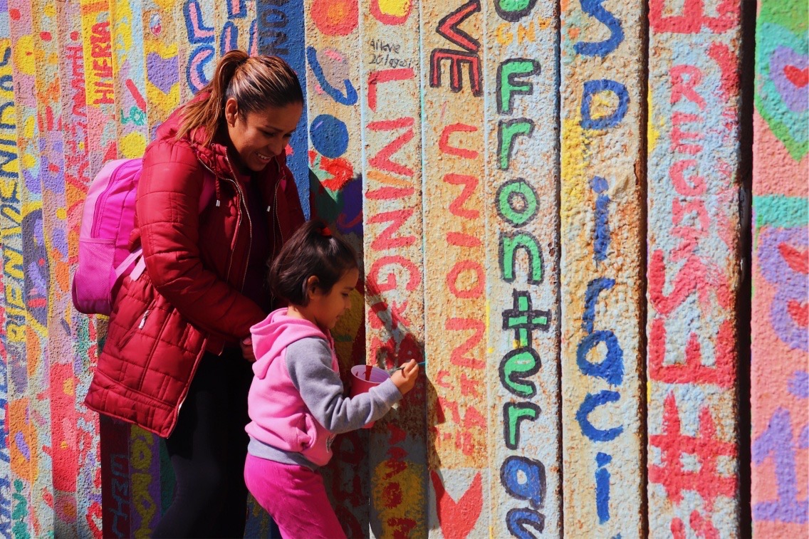 Photograph of a mother wearing a red puffer coat and her young daughter's school backpack, standing in front of a ridged mural wall with her daughter, wearing pink. The daughter uses red paint from a red Solo cup to paint on the wall.