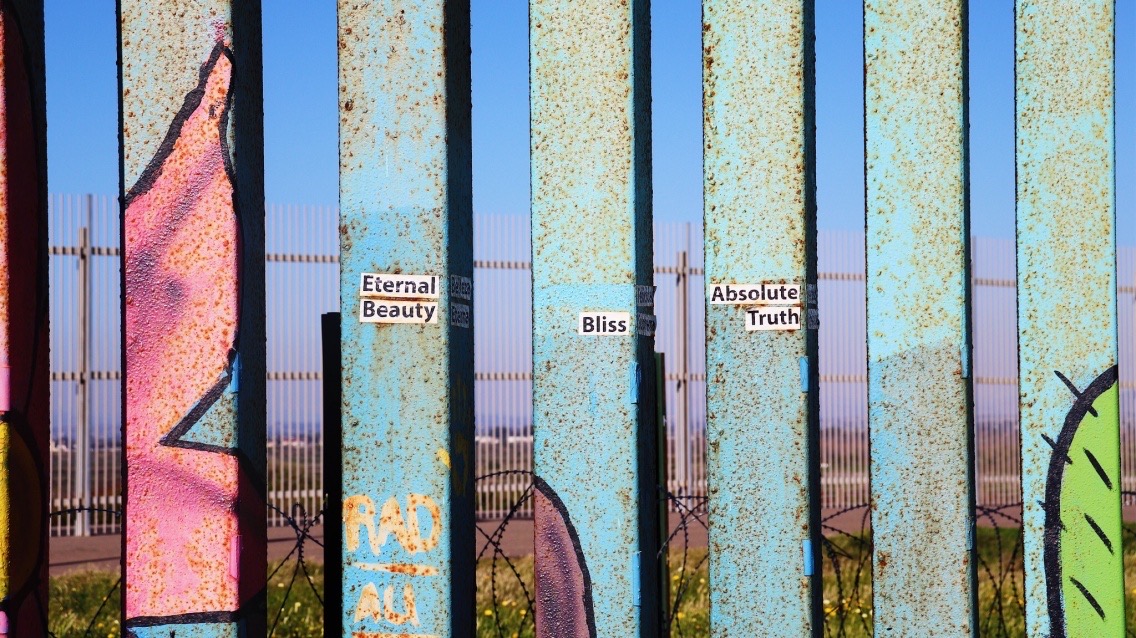 Photograph of 6 metal columns, painted in blue, pink and green. The middle three columns have stickers with words: 'eternal beauty', 'bliss', 'absolute truth' (from left to right). Behind the columns is a blue sky and two fences: one with skinny metal poles, the other a circular barbed wire fence.