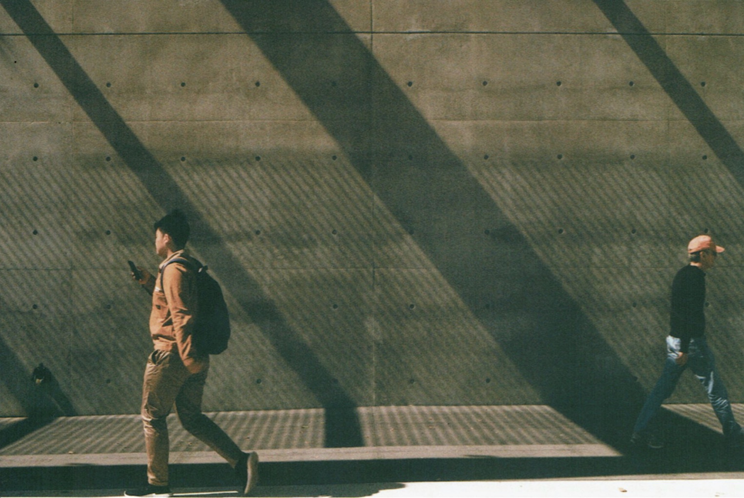 Medium: Film Photography. Light shines through slits to form a trapezoidal distortion against a concrete wall with drill holes. A student walks left while reading from his phone, while a man on the right moves in the other direction.