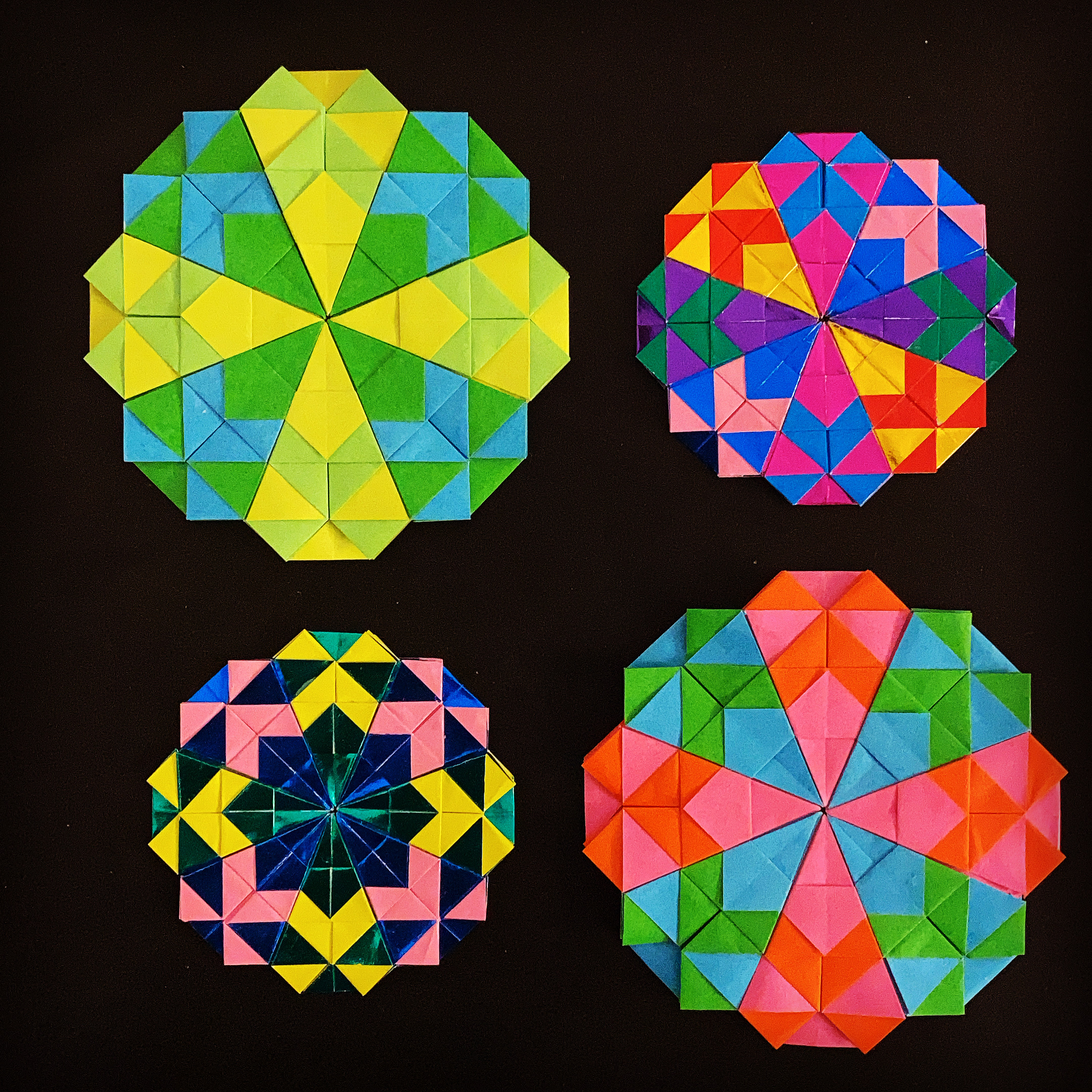 Four different color variations of the same flower origami layout. There are eight leaves in each flower. Top-left: lime green and yellow alternate with the green and blue leaves; top-right: an assortment of strong colors in each leaf; bottom-left: an assortment of darker colors in each leaf; bottom-right: orange and pink alternate with green and blue leaves.