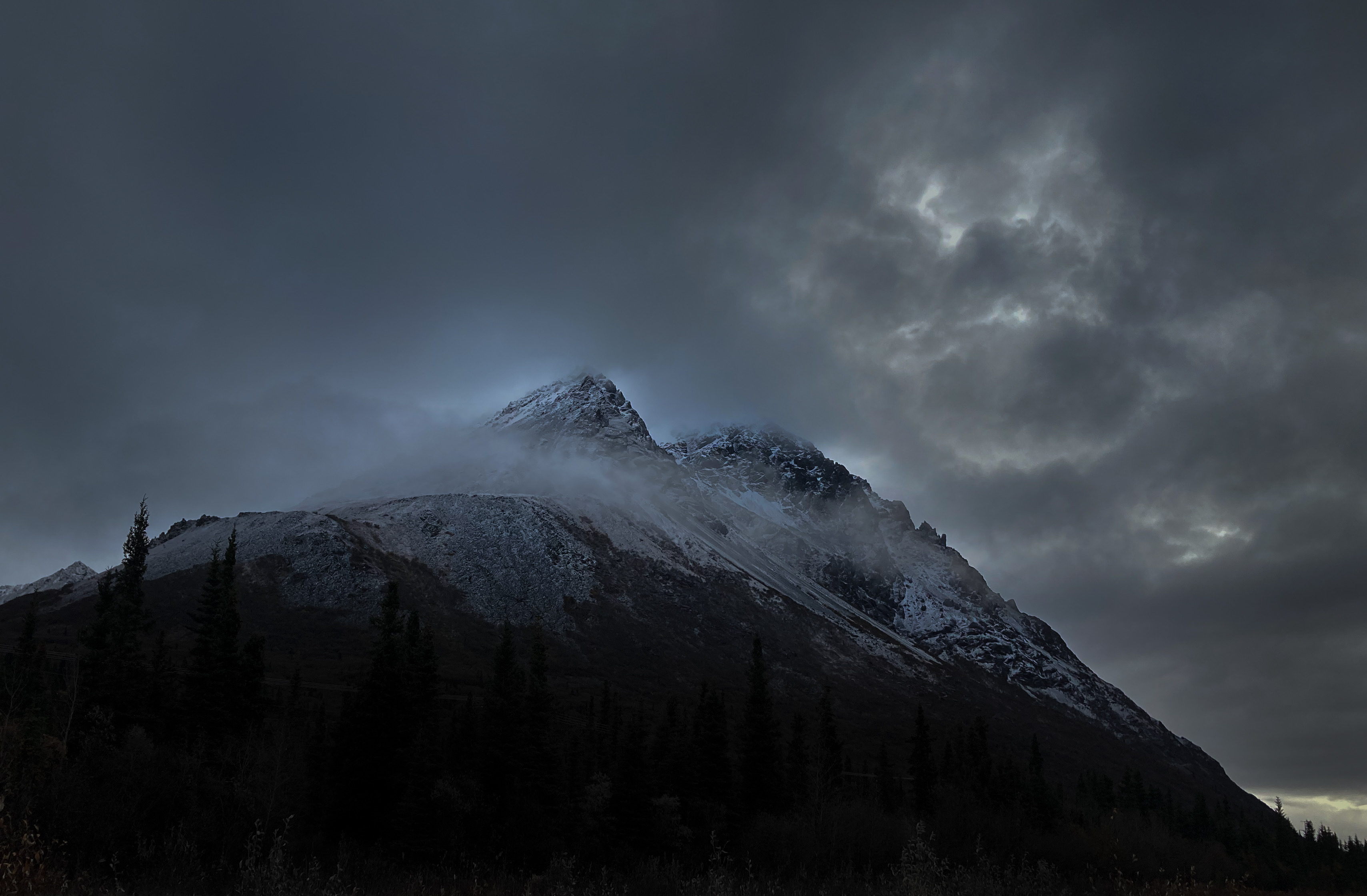 Photograph of a snow-topped mountain on a dark, cloudy day. Small pockets of light can be seen between clouds.