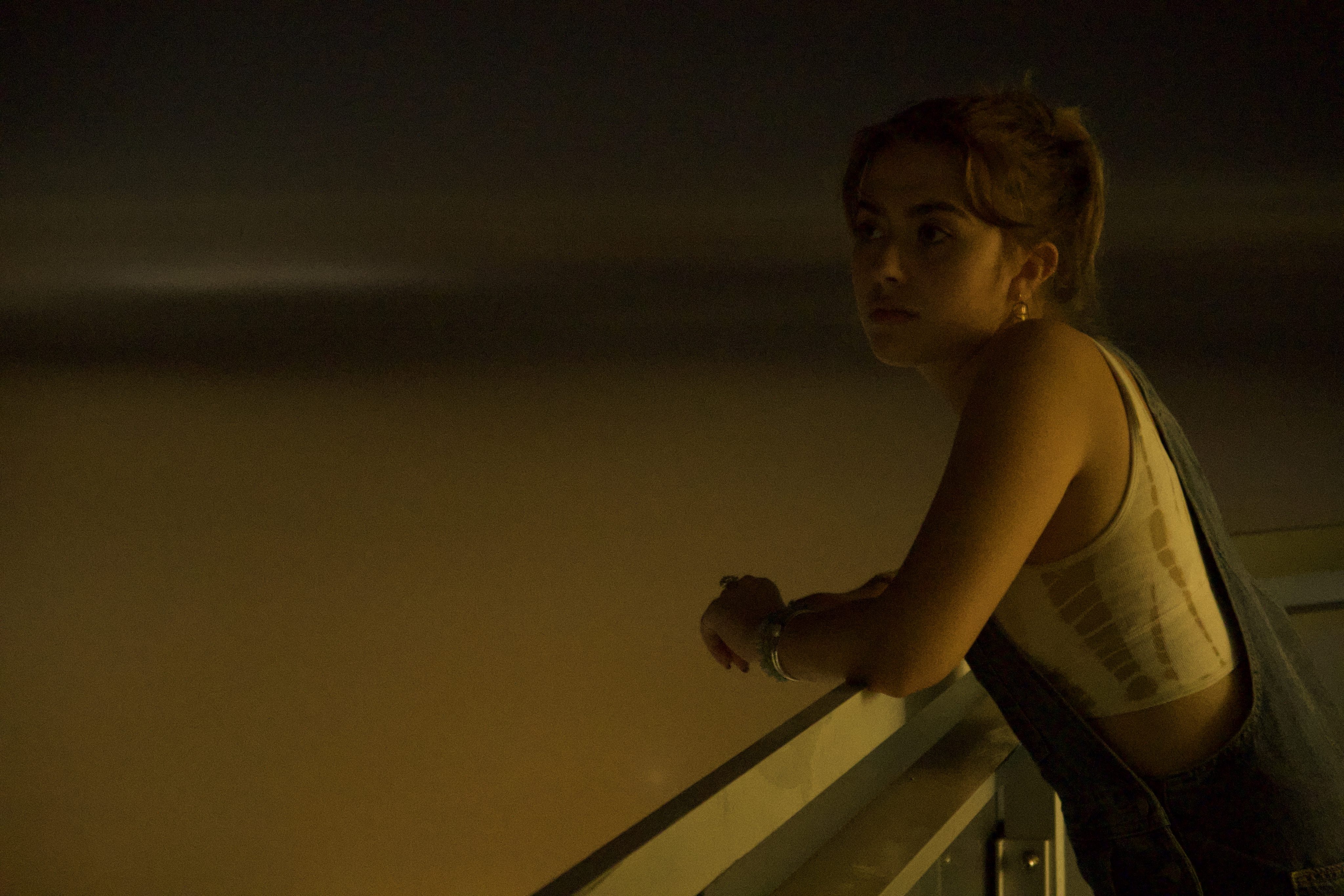 Photograph of a girl in a tank top and overalls leans over a railing, looking off into the distance.